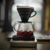 A pour-over coffee setup with a black dripper over a glass carafe filled with dark coffee. The scene has a cozy, relaxed atmosphere.