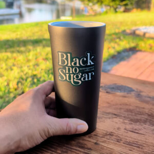 A hand holds a black travel mug on a wooden table outdoors; the mug reads "Black no Sugar" in large letters. Grass and water are visible in the background.
