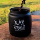 A black coffee canister labeled "Black No Sugar" on a wooden table, with coffee beans being poured in. Background shows green grass, suggesting an outdoor setting.