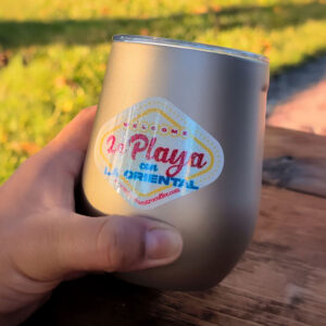 A hand holds a silver cup with a colorful logo reading "La Playa con La Oriental." The cup rests on a wooden surface with grass in the background, conveying a relaxed, outdoor vibe.
