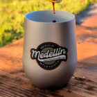 Close-up of a metal mug labeled "Brewed in Medellin," with coffee being poured in. The scene is set outdoors on a sunny day, creating a warm, inviting tone.