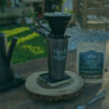 A serene outdoor setting with a coffee setup on a wooden table. A black kettle, mug, and pour-over dripper are in focus beside a bag of coffee beans.