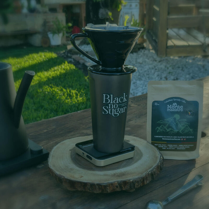 A serene outdoor setting with a coffee setup on a wooden table. A black kettle, mug, and pour-over dripper are in focus beside a bag of coffee beans.