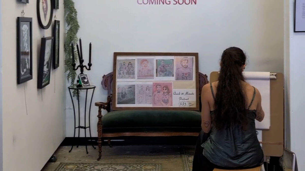 A person with long hair, seated and drawing. They face a display of portraits on a couch. The white wall above reads "COMING SOON," evoking anticipation.