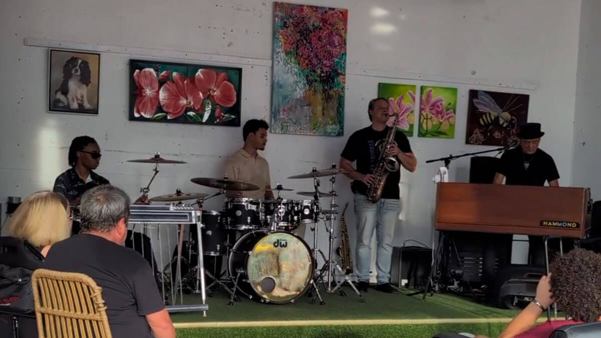 A jazz band performs in an art-filled gallery. A saxophonist, drummer, keyboardist, and another musician create a lively, relaxed atmosphere.