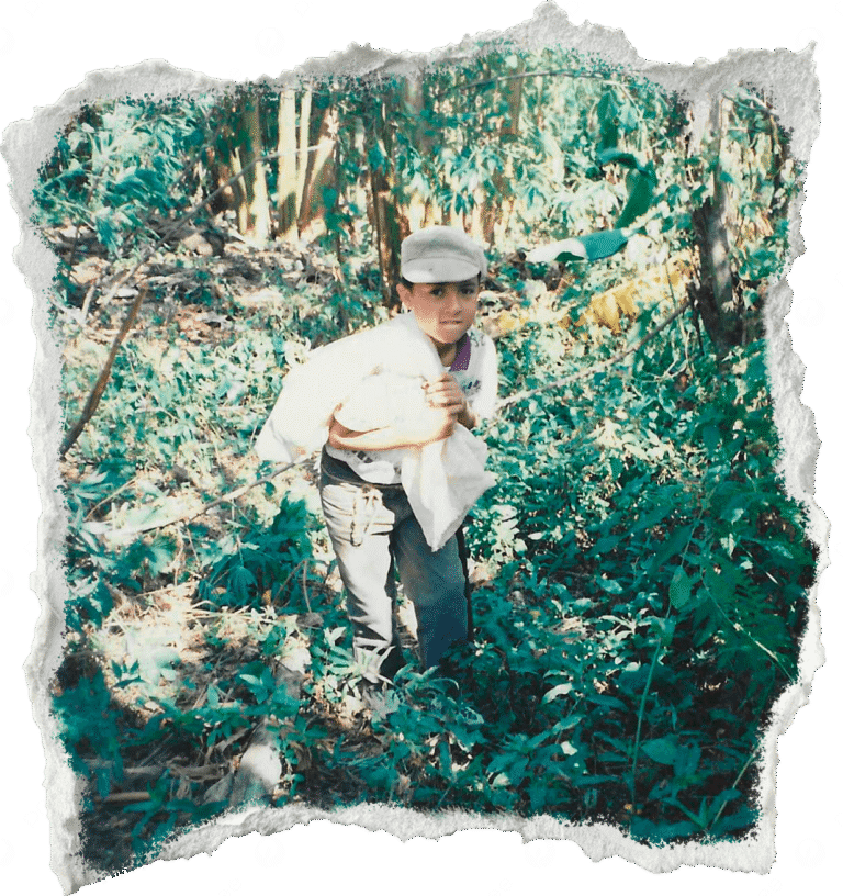A boy in a cap and casual clothes is in a lush, leafy forest, carrying a white sack. The photo has rough, torn edges, conveying a vintage feel.